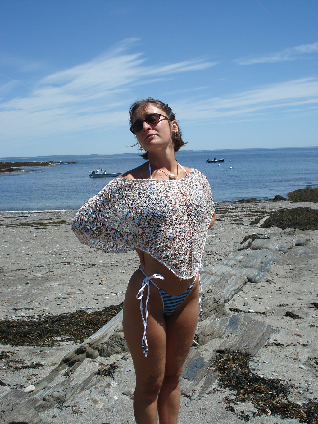 Woman in a knit poncho and blue bikini standing on a rocky beach with ocean and sky in the background.