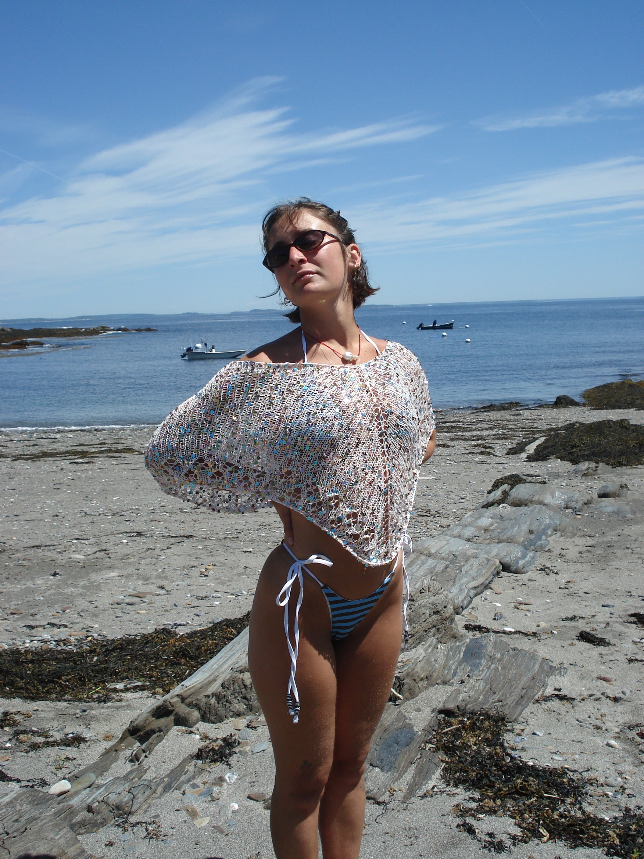 Woman in a knit poncho and blue bikini standing on a rocky beach with ocean and sky in the background.