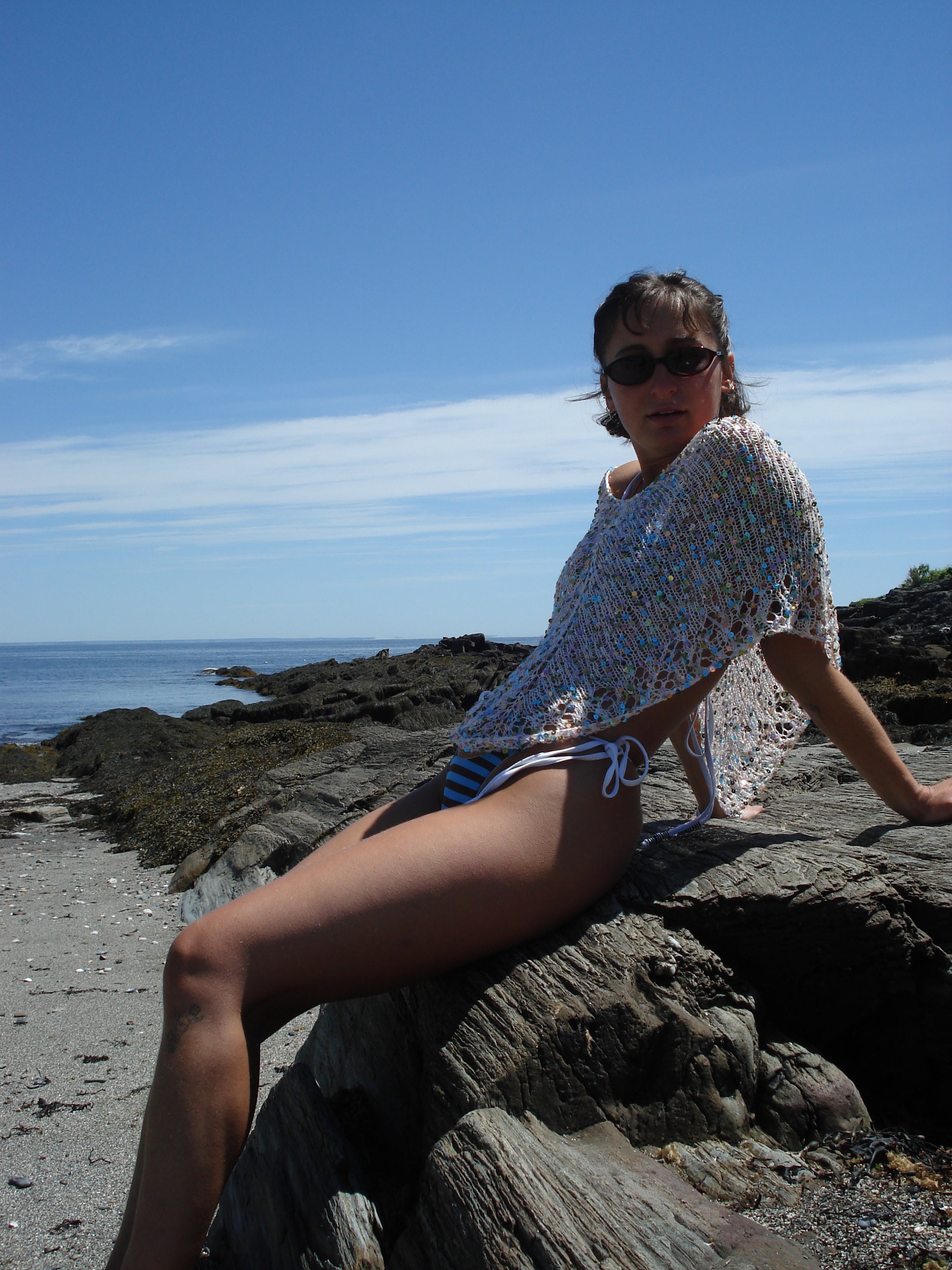 Woman sitting on a rock by the ocean wearing sunglasses and a knit poncho.